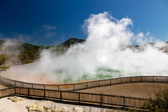 View of Waiotapu Thermal Wonderland with steam rising from the geothermal pool, surrounded by a wooden fence under a clear blue sky in Rotorua, Waikato Region, New Zealand.