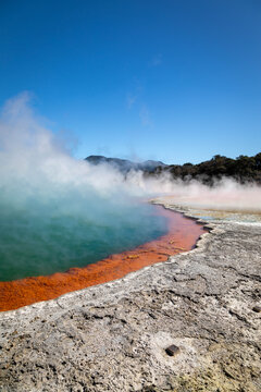 View of the Waiotapu Thermal Wonderland geothermal pool with vibrant orange mineral deposits and rising steam under a clear blue sky in Rotorua, Waikato Region, New Zealand.