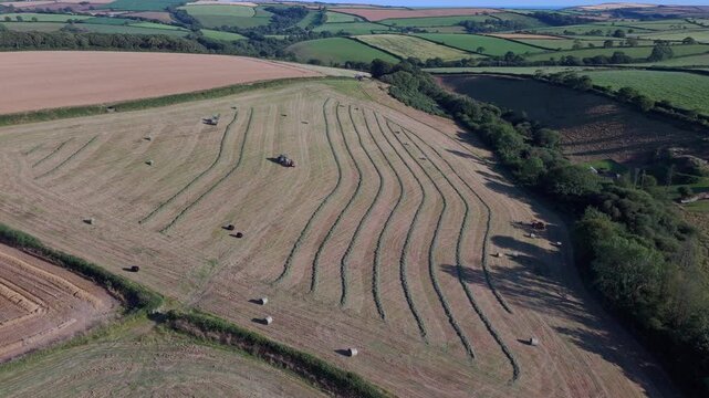 Aerial view of tractors harvesting hay and wrapping bales in the summer sun across the rural landscape of Looe, England, United Kingdom.