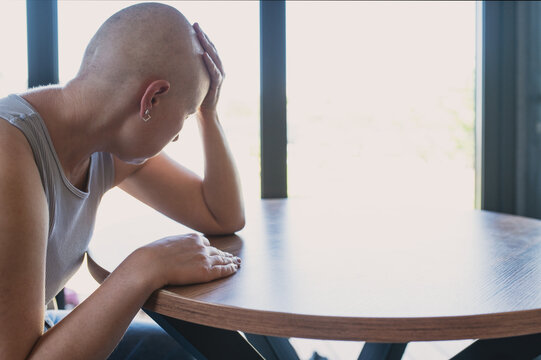 A bald woman sits by the window, resting her head on her hand. Sadness, depression, loneliness. Coming to terms with a cancer diagnosis.