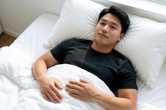 Young Asian man lies on white bed after waking up. Sleepy male wearing black t-shirt rests on pillow in bedroom. Tired guy looks at camera relaxing at home in morning. Healthy lifestyle, calm sleep