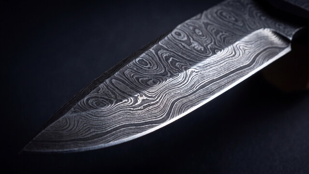 Close up view of a sharp damascus steel knife blade with intricate wavy patterns resting on a dark textured background surface