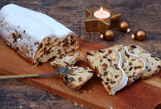 Traditioneller deutscher Christstollen mit Puderzucker auf Holzbrett, aufgeschnitten in Scheiben mit Weihnachtsdekoration und Kerze.