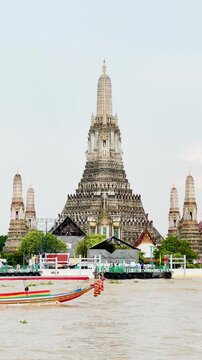 Wat Arun temple rises beside the Chao Phraya River in Bangkok, Thailand, as a long-tail boat cruises past during a peaceful evening with soft clouds and warm sunset light over the riverside.