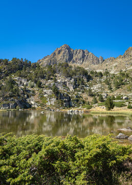 View of Llac de Pessons surrounded by rocky mountain peaks, pine trees, and clear blue sky in Grau Roig, Encamp, Andorra.