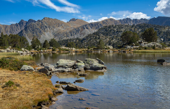 View of Llac de Pessons surrounded by rocky shores, pine trees, and rugged mountain peaks under a blue sky in Grau Roig, Encamp, Andorra.