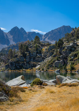 View of Llac de Pessons surrounded by rocky terrain, pine trees, and mountain peaks under a clear blue sky in Grau Roig, Encamp, Andorra.