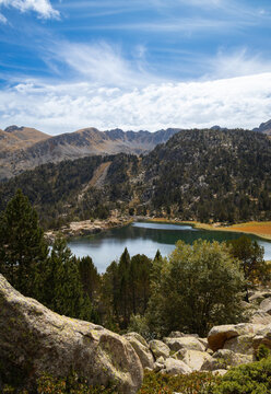 View of Llac de Pessons surrounded by pine trees, rocky terrain, and mountains under a blue sky with clouds in Grau Roig, Encamp, Andorra.