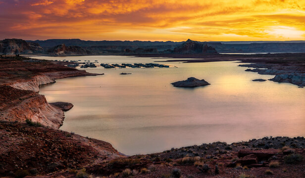 View of Lake Powell at sunrise with low water levels, exposed rocky shoreline, boats, and sandstone formations in the drought-affected Colorado River basin.