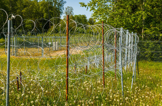 View of a border fence with razor wire, green grass, and dandelions in a field in Europe.