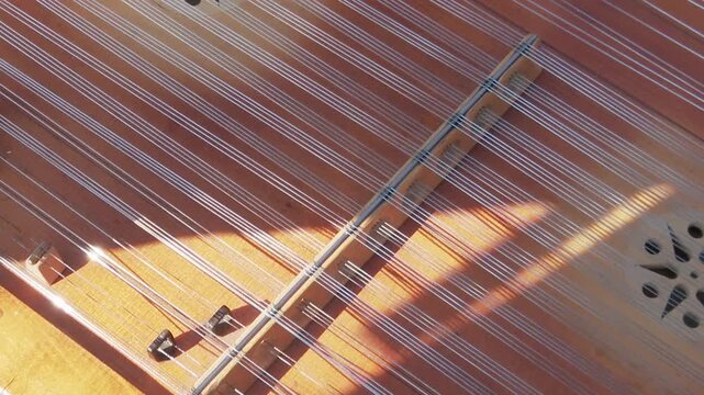 A close-up shot of the strings and wooden soundboard of a cimbalom. Sunlight creates bright flare and reflections
 on the vibrating metal strings during a musical performance.