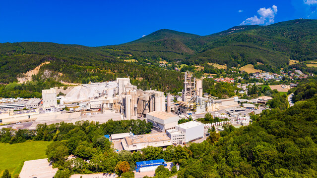 Cement towers and warehouses occupy a mountain valley.. Gray silos and pale roofs occupy an Austrian industrial site below green hills and scattered houses.