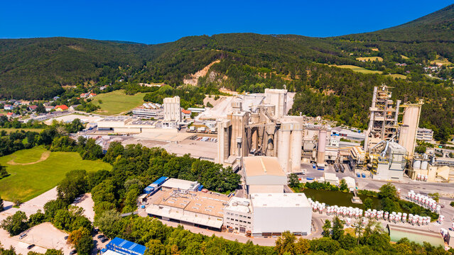 A silo cluster rises above warehouse roofs at a cement plant.. Gray towers stand behind tan and white roofs, with dense green trees along the site boundary.