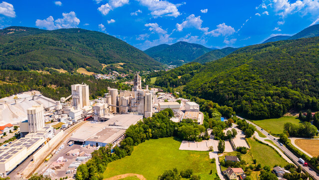 A cement production complex stands below mountain slopes.. Gray towers at an Austrian cement plant border a green meadow beneath wooded slopes and white clouds.