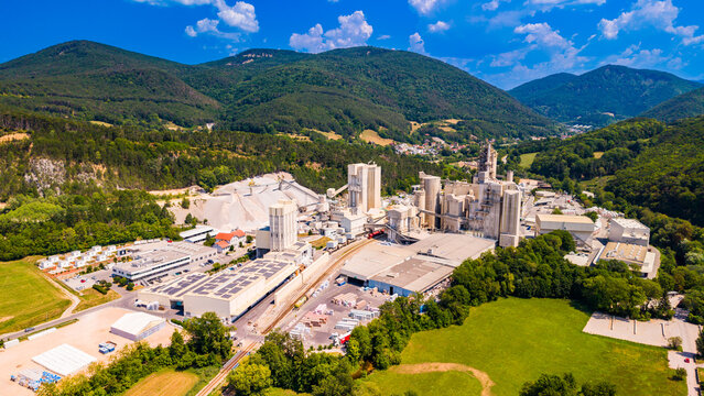 Cement warehouses and silos stand between farmland and forest.. Pale roofs and gray towers occupy a valley site framed by green fields and wooded slopes.