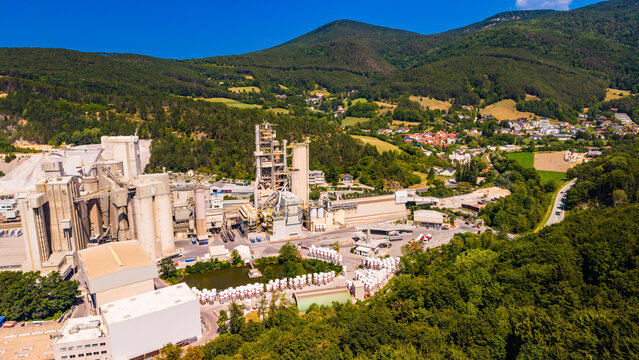 A road curves through a valley beside cement storage structures.. A road curves past gray silos and pale buildings below green hills and scattered houses.