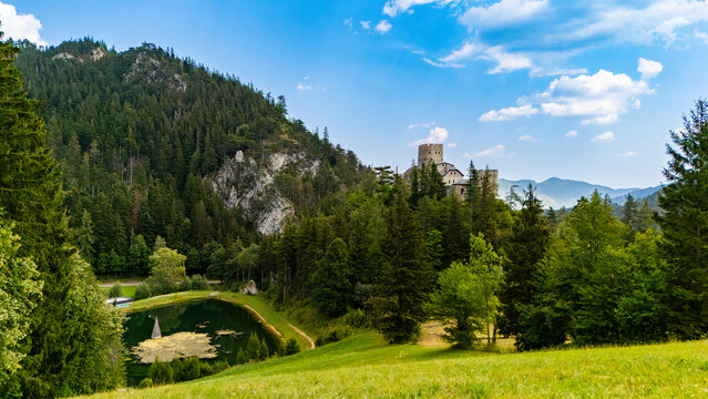 A stone castle stands beside a pond below a rocky hillside.. A pale Austrian castle tower rises behind green trees beside a dark pond and sunlit grass.
