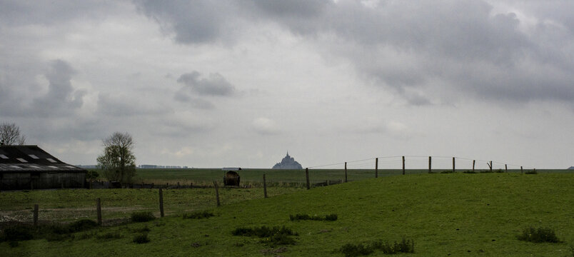 View of Mont Saint-Michel in the distance across green fields with a wooden fence and a rustic barn under a cloudy sky in Mont Saint-Michel, Normandy, France.