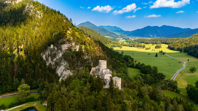 A stone castle stands beside a wooded cliff above valley fields.. Pale Austrian castle towers stand among green trees beside exposed rock, with a pond below.