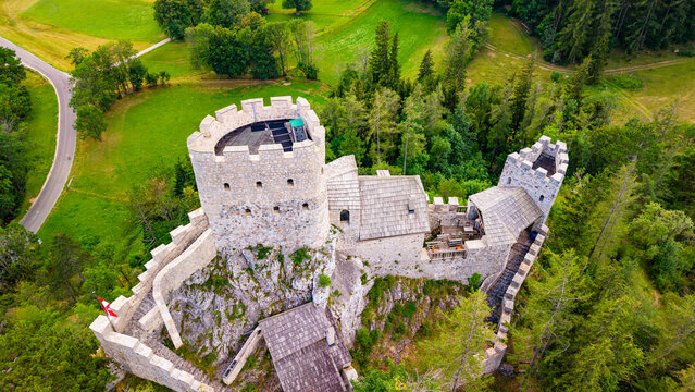 An Austrian stone castle encloses a courtyard around its tower.. Pale battlements surround a tower and tiled roofs on exposed rock beside green grass.