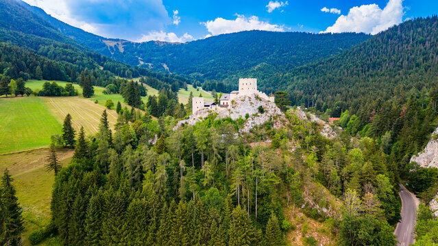 Stone towers rise above forest and fields at a hilltop castle.. Pale walls of an Austrian castle crown a rocky outcrop amid green conifers, with open farmland behind.