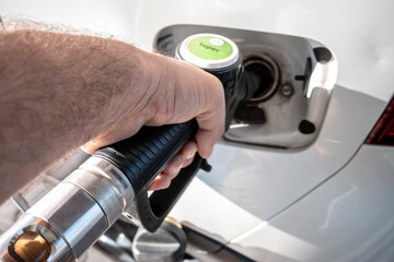 A close-up of a person’s hand holding a fuel nozzle as they refuel a white car, focusing on the pump, gas cap and nozzle illustrating refueling, energy, travel and car maintenance. © simona