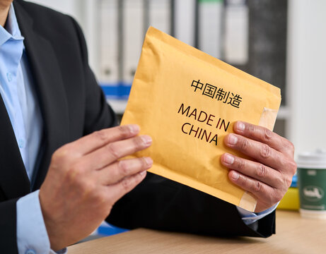 Close-up of a businesswoman's hands opening a package labeled "Made in China" in Chinese characters, while sitting at a desk in her home.