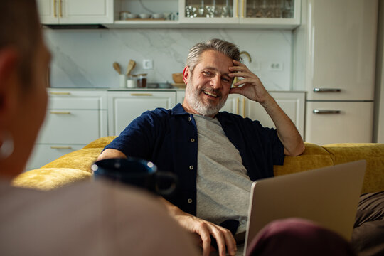 Smiling middle-aged man chatting on sofa with laptop at home