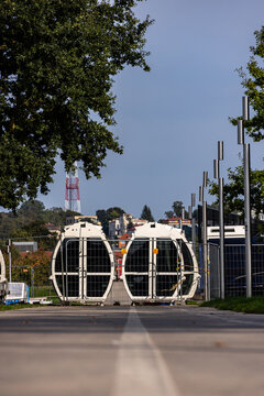 View of two white cable car cabins resting on a paved path behind a metal fence with trees and a radio tower in the background in Kaunas, Kaunas County, Lithuania.