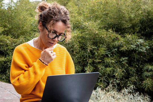 Woman in yellow sweater focuses on laptop while working remotely outdoors, thoughtful as she reads, researches and manages tasks in a peaceful garden with green foliage and plants.