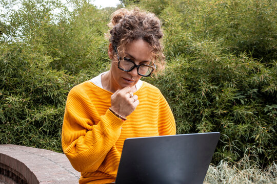 Thoughtful woman in a yellow sweater using a laptop outdoors doing remote work, enjoying nature while studying notes and making decisions with calm focus and quiet determination.