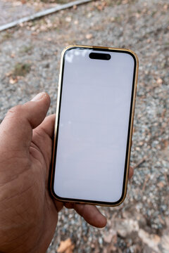 Hand holding smartphone with blank white screen outdoors over gravel ground, close-up of modern mobile device in a gold case, ideal for app mockup, communication and mobile tech.