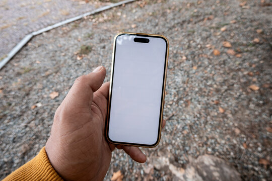 Hand holding smartphone with a blank white screen outdoors over a gravel path with scattered autumn leaves, close-up perspective emphasizing the device and hand in natural light