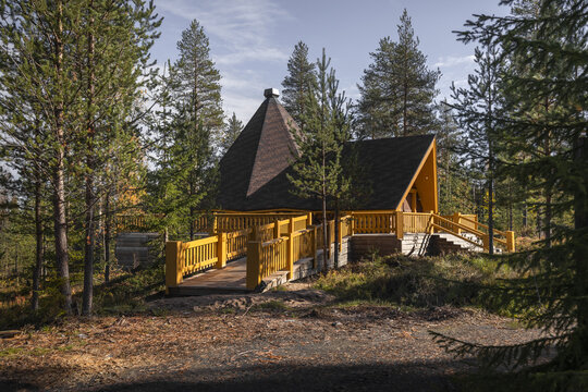 View of a traditional wooden hut with a pointed roof and a wooden walkway nestled among pine trees in the forest in Rovaniemi, Lapland, Finland.