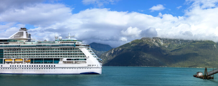 View of a large cruise ship docked in the waters of a fjord with dramatic mountains and a cloudy sky in the background in Haines, Alaska, United States.
