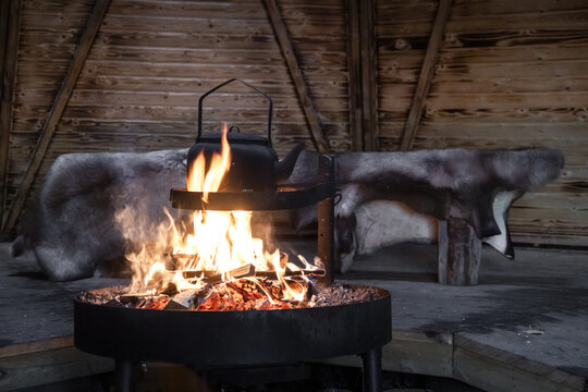 View of a crackling fire pit with a black kettle inside a traditional wooden hut with reindeer hides in the background at Apukka Resort in Rovaniemi, Lappi, Finland.