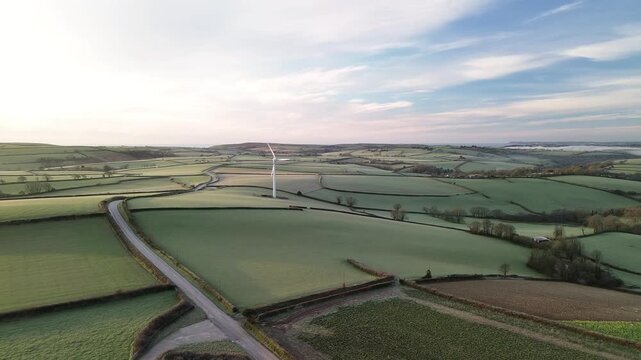 Aerial view of a wind turbine generating sustainable energy in a frosty rural landscape with hedgerows and a country road at sunrise in Looe, England, United Kingdom.
