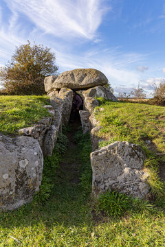 View of the ancient Sprovedyssen dolmen with large megalithic stones, grassy mounds, and a person standing in the entrance under a blue sky in Stege, Mon, Denmark.