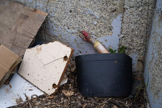 View of a discarded doll arm protruding from a black plastic container amidst scattered debris, cardboard, and broken tiles in Bulgaria.