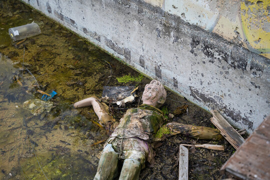 View of a weathered and damaged mannequin lying in shallow, murky water against a textured concrete wall in Bulgaria.
