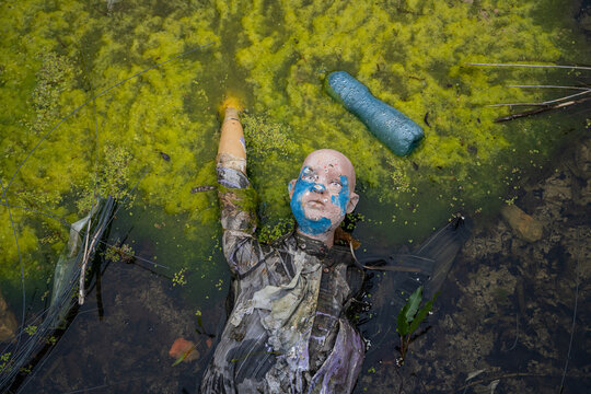 View of a discarded mannequin partially submerged in a pond covered with green algae and duckweed with a plastic bottle floating nearby in Bulgaria.
