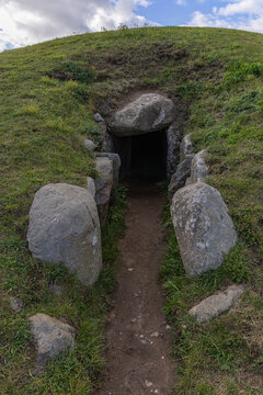 View of the entrance to the Kong Asgers Hoj Neolithic passage grave, featuring large stone slabs set into a grassy mound under a cloudy sky in Stege, Mon, Denmark.