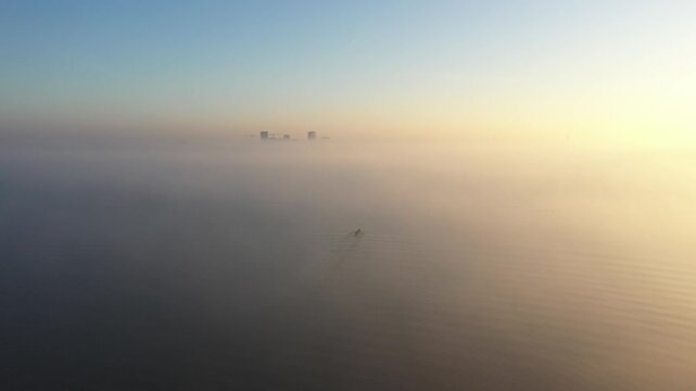 Aerial view of a lone rower in a small boat gliding across calm water shrouded in thick morning fog near the city skyline in Almere, Flevoland, Netherlands.