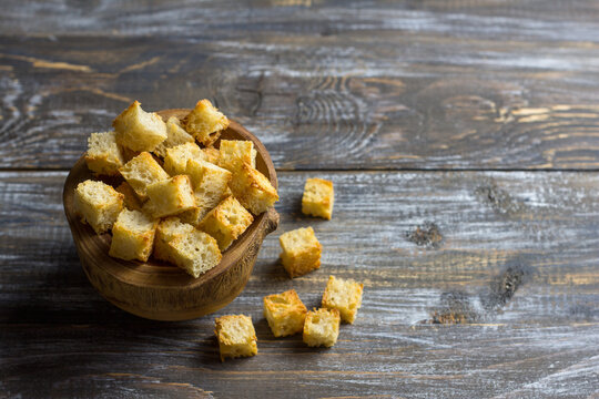 Golden homemade croutons in a rustic wooden bowl on weathered dark wood with scattered toasted bread cubes, warm light, copy space