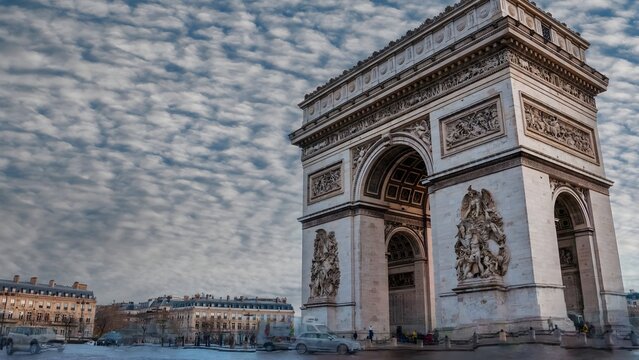 View of the Arc de Triomphe with its intricate stone carvings under a dramatic cloudy sky in Paris, Ile-de-France, France.