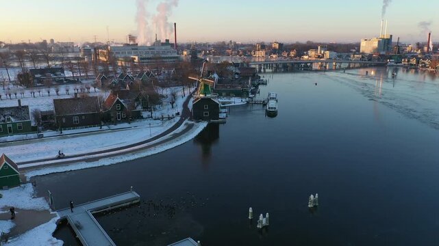 Aerial view of the historic Zaanse Schans windmills along the Zaan river with snow-covered banks and industrial buildings in the background in Zaandam, Noord-Holland, Netherlands.