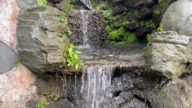 Small artificial garden waterfall flowing over mossy stones in a tranquil outdoor setting.