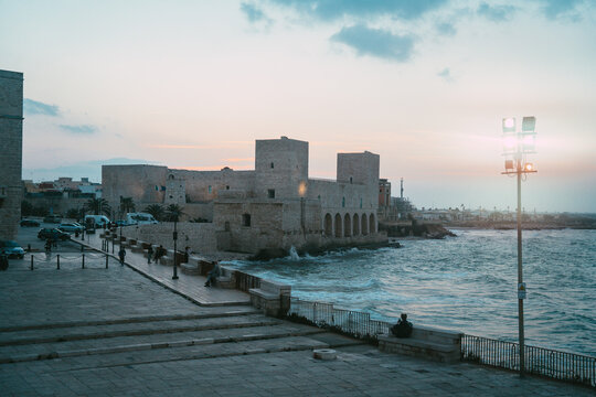 View of the historic Swabian Castle of Trani situated along the rocky coastline with waves crashing against stone walls and people on the promenade at dusk in Trani, Apulia, Italy.
