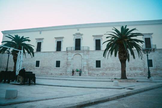 View of the historic stone facade of a building with palm trees and people standing in the square in Trani, Apulia, Italy.