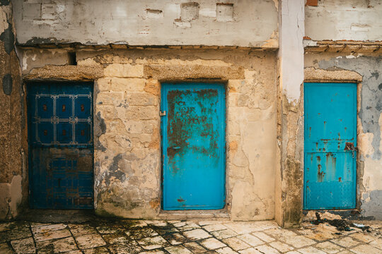 View of three weathered blue and turquoise doors set into a rustic stone wall with peeling plaster and cobblestone ground in Bari, Apulia, Italy.